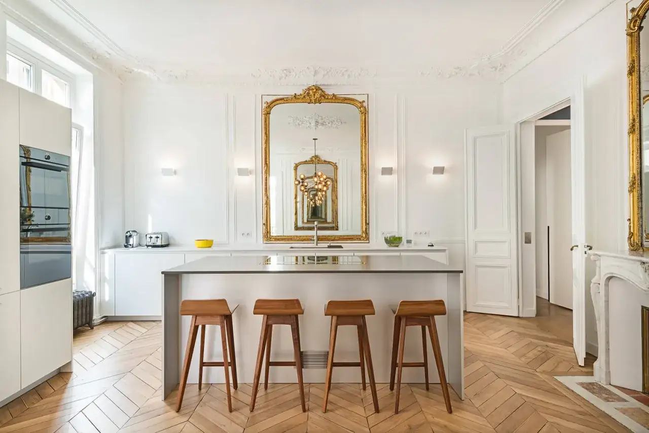 Cuisine lumineuse au style classique, avec un grand miroir doré, un îlot central et des tabourets en bois sur un parquet en chevrons.