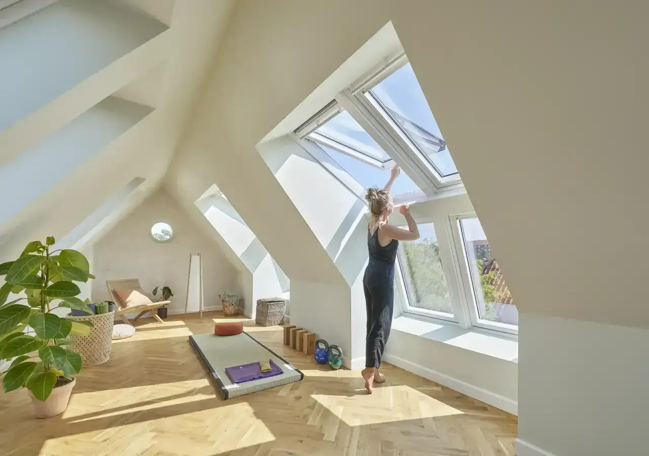 Frau &ouml;ffnet Dachfenster im ausgebauten Dachboden, der als Yoga-Raum eingerichtet ist. Viel Licht und Holzfu&szlig;boden.