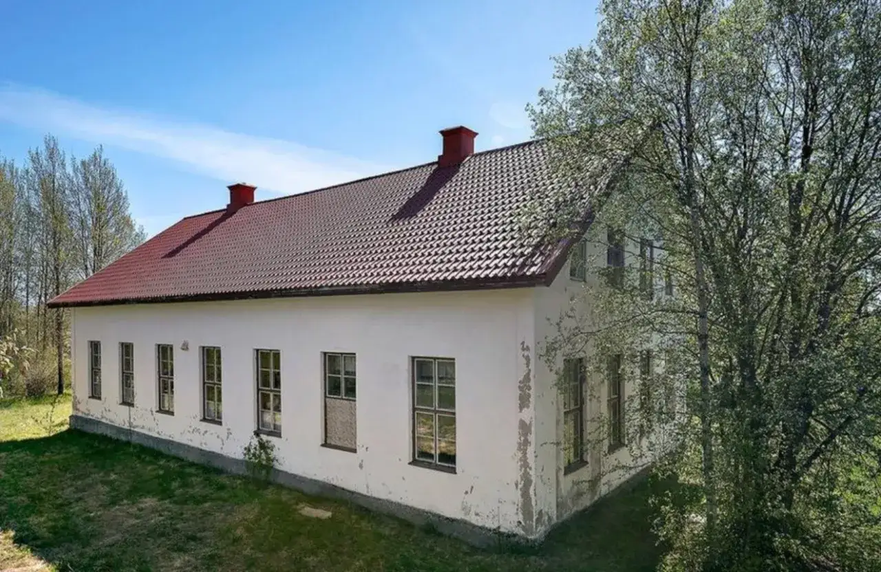 Vita gamla hus med rött tegeltak och skorstenar, omgivna av träd och gräs under en blå himmel.