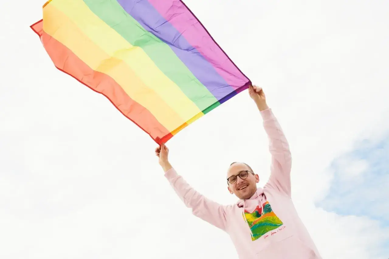 Uśmiechnięty mężczyzna z okularami trzyma tęczową flagę, świętując kiedy jest dzień LGBT.