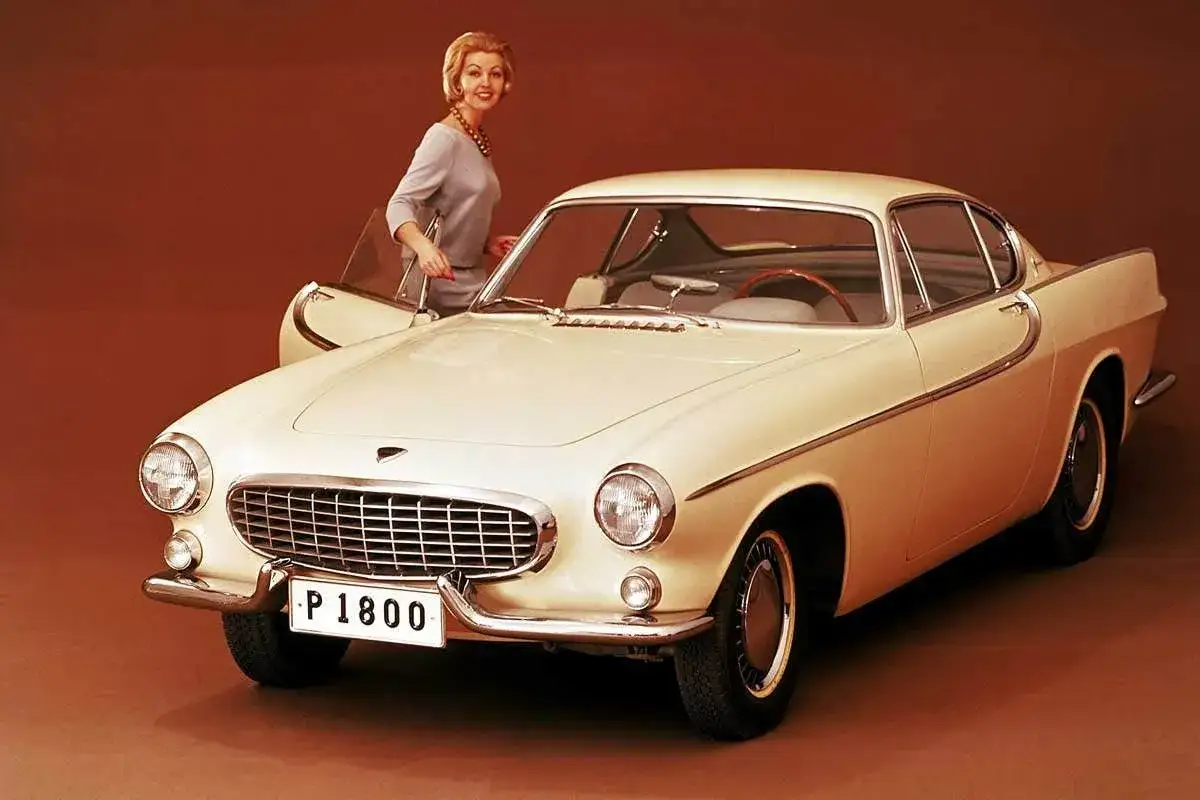 En kvinna med kort blont hår och en ljusblå klänning lutar sig mot en klassisk, ljusgul Volvo P1800. En tidlös skönhet att sugga bil.