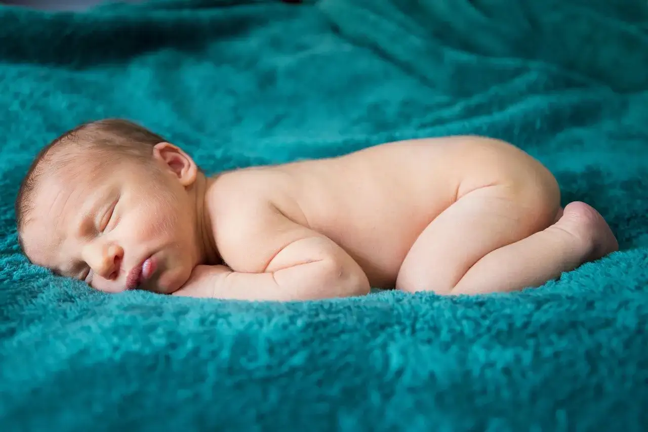 Neonato addormentato su plaid turchese, con una piccola fossetta sacrale visibile.