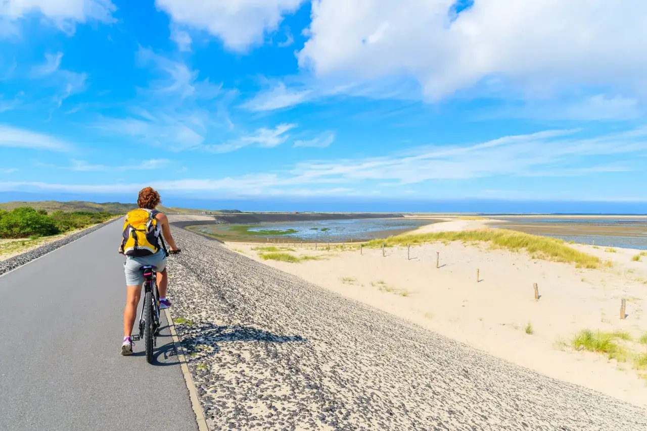 A woman enjoys a bike trip in Denmark, cycling along a coastal path with sand dunes and the sea under a bright blue sky.