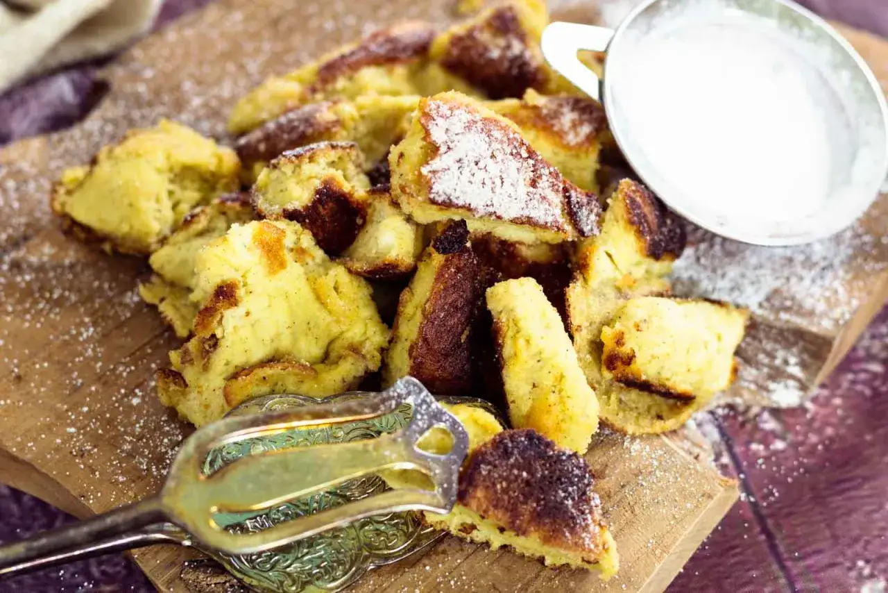 Ein Haufen goldbrauner, zerrissener Stücke von **low carb Kaiserschmarrn**, bestäubt mit Puderzucker. Daneben ein kleines Schälchen mit Puderzucker und eine elegante Gabel.