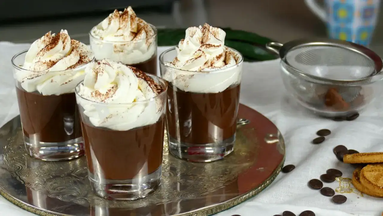 Quattro bicchierini di cremoso budino al cioccolato, sormontati da panna montata e spolverati di cacao. Un dolce invitante.
