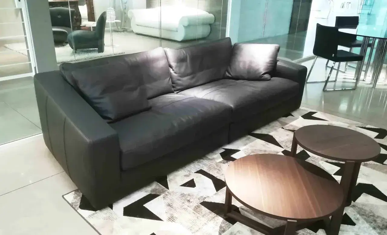 Un elegante divano angolare in pelle nera, con due tavolini rotondi in legno, su un tappeto geometrico. Un frighetto &egrave; visibile in lontananza.
