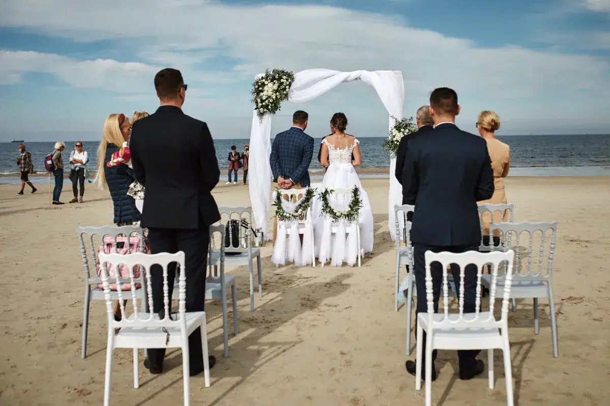 Uroczysty ślub na plaży. Para młoda stoi przed białym baldachimem, goście obserwują ceremonię.