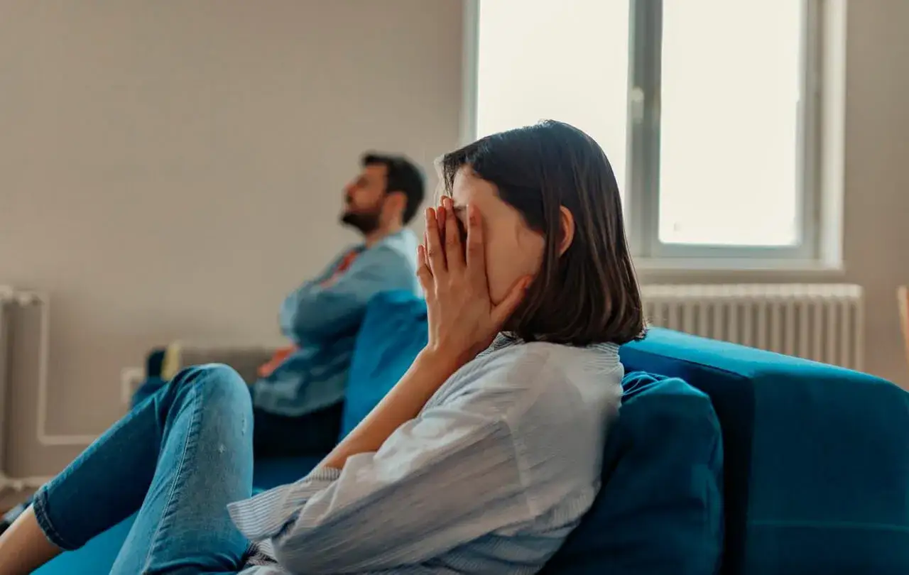 Un couple assis dos à dos sur un canapé bleu, visiblement en conflit. La femme se cache le visage dans les mains, suggérant un profond désarroi, peut-être lié à un manque sexuel.