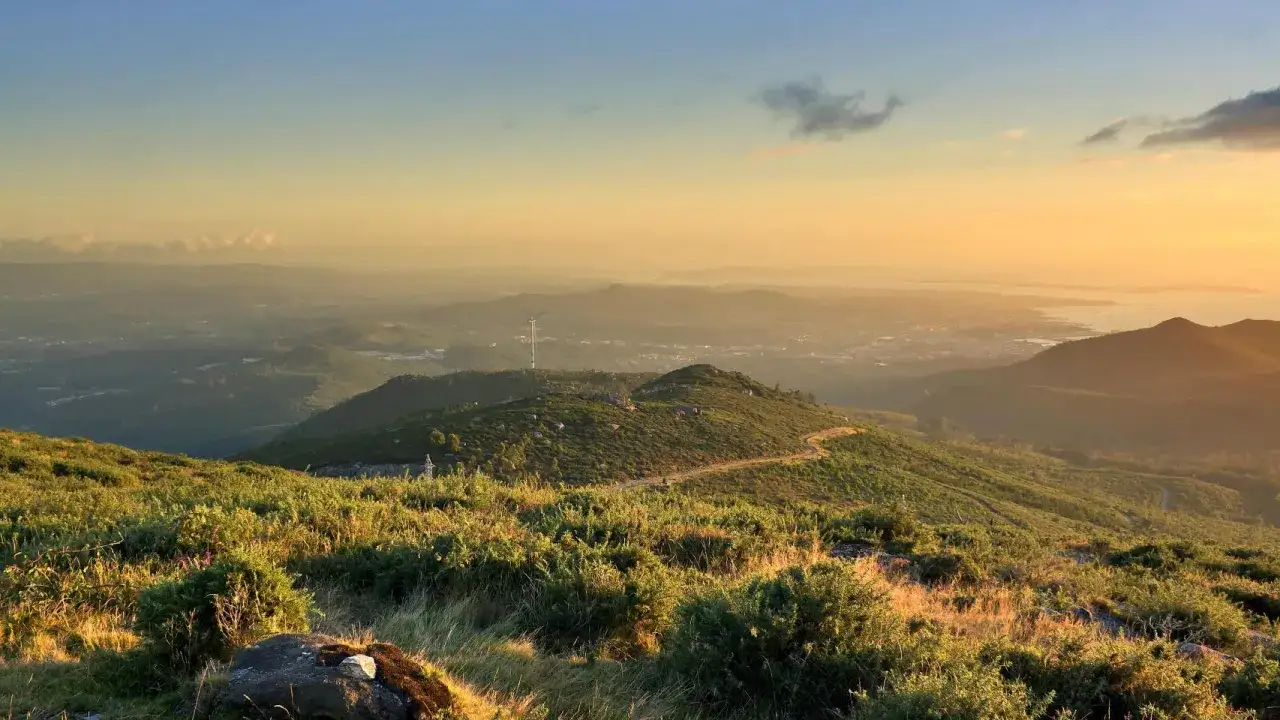 Atardecer dorado sobre el monte Xiabre, con un aerogenerador solitario y un camino serpenteante entre colinas verdes.