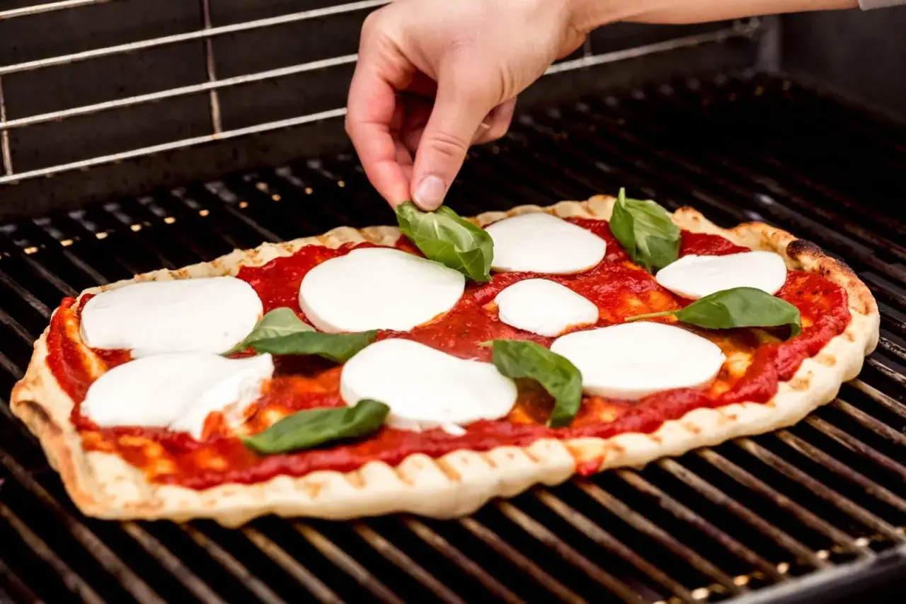 Ostatnie szlify na pizzy z grilla gazowego. Dłonie dodają świeżą bazylię na mozzarellę i sos pomidorowy.