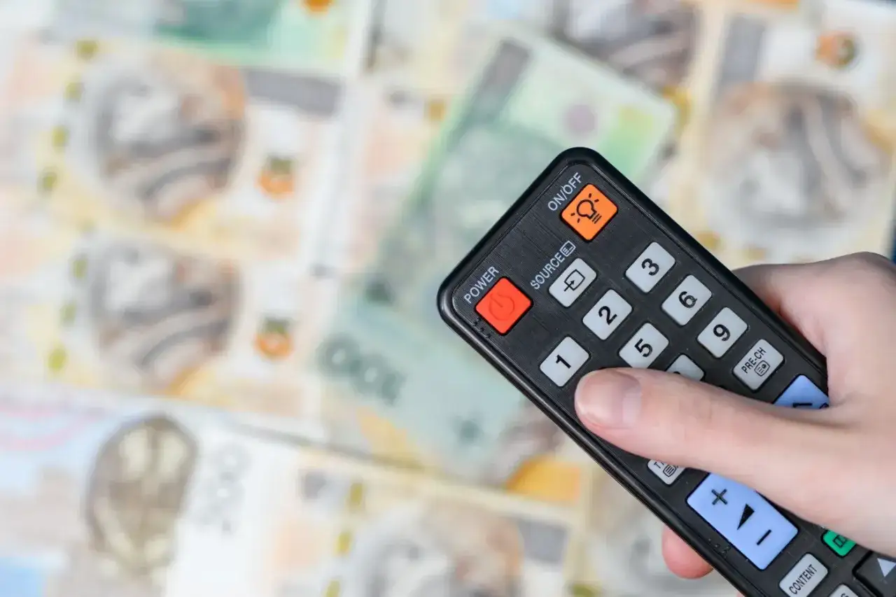 Dłoń trzymająca pilot do telewizora nad stosem polskich banknotów. Symbolizuje to likwidację abonamentu RTV i oszczędności.
