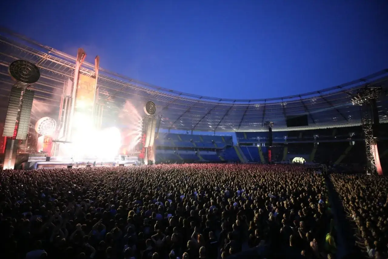 Ogromny tłum na stadionie podczas koncertu Rammstein. Scena rozświetlona jest potężnymi światłami i pirotechniką.