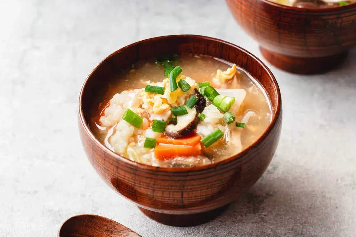 A steaming bowl of Japanese rice soup, filled with tender vegetables, mushrooms, and scrambled egg, garnished with fresh green onions.