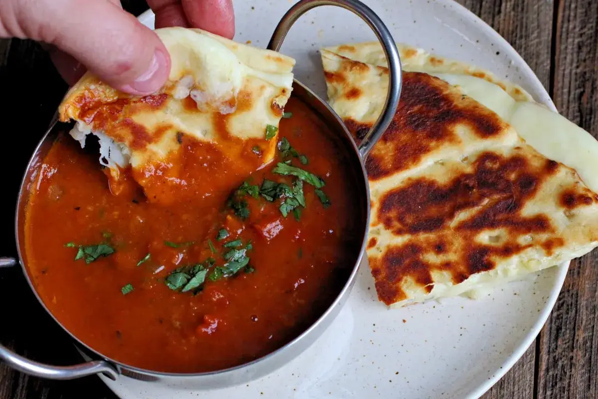 Ein St&uuml;ck Naan-Brot wird in eine Sch&uuml;ssel mit w&uuml;rziger, roter Masala-Sauce getunkt, garniert mit frischen Kr&auml;utern.
