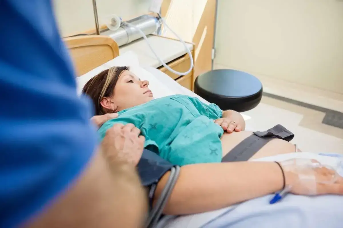 Kobieta leży w łóżku szpitalnym, z ręką lekarza na brzuchu. Monitoruje się jej stan po porodzie, zwracając uwagę na nieprawidłowe obkurczanie macicy.