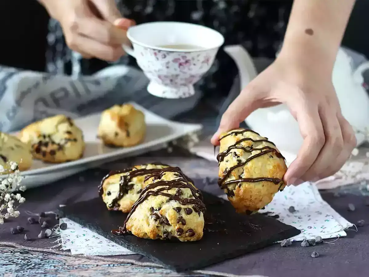 En hand plockar upp en av de läckra scones med choklad, doppade i mörk choklad. En kopp te står redo.