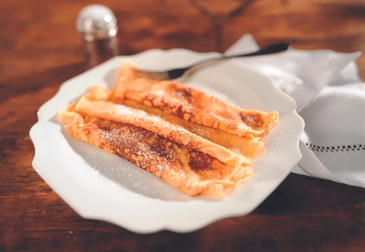 Drei goldbraune Palatschinken, mit Puderzucker bestreut, auf einem weißen Teller. Ein perfektes Palatschinken Rezept für jeden Anlass.