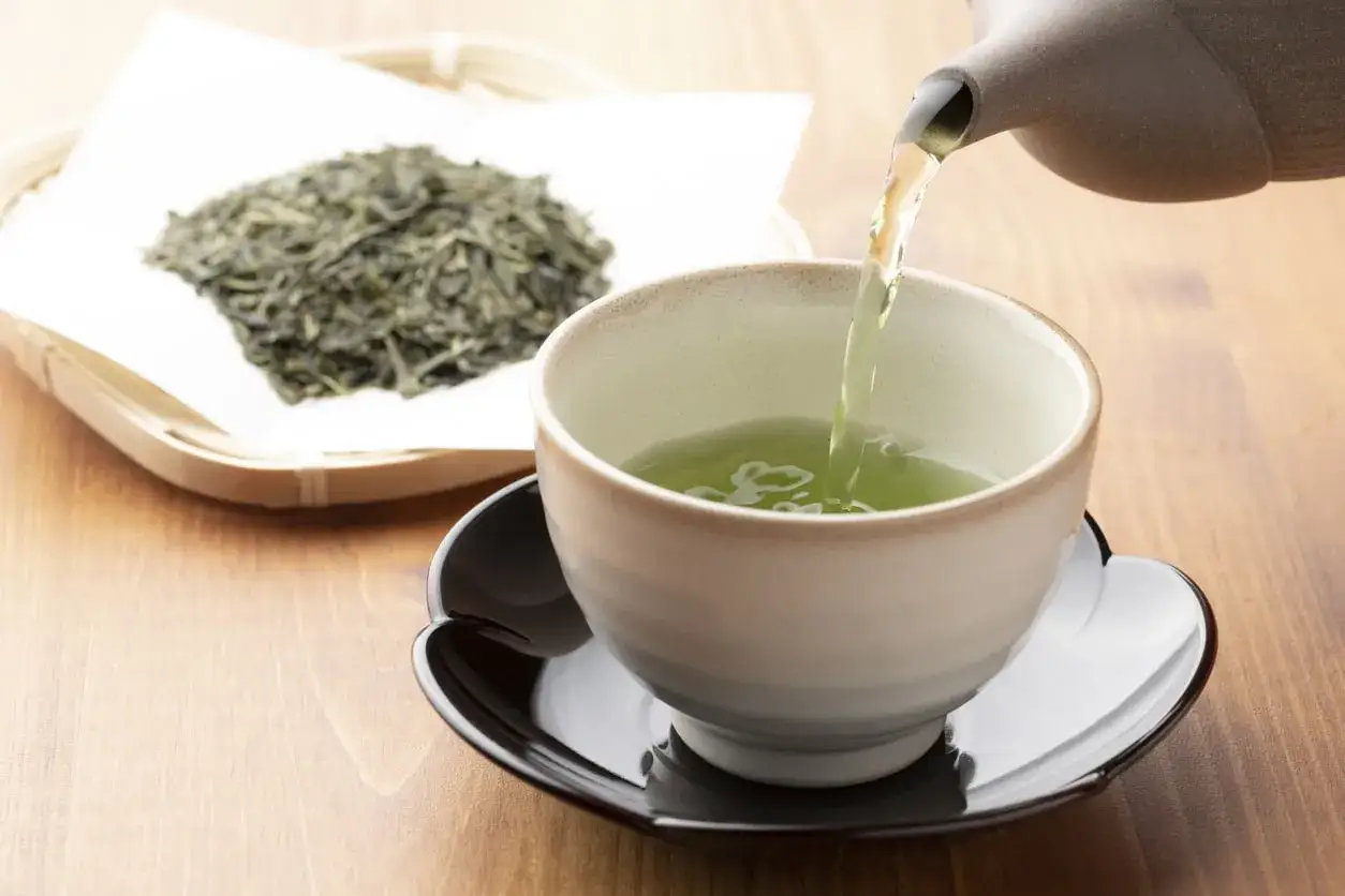 Versando tè verde in una tazza, gustando i suoi benefici per la salute. Foglie di tè secche in un cestino sullo sfondo.