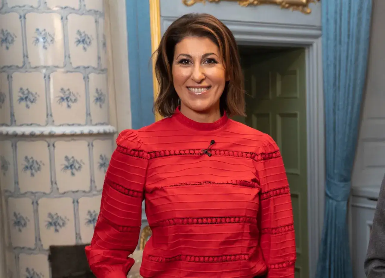 Rania Shemoun Olsson ler glatt i en röd topp med puffärmar. Bakom henne syns en kakelugn och blåa draperier.