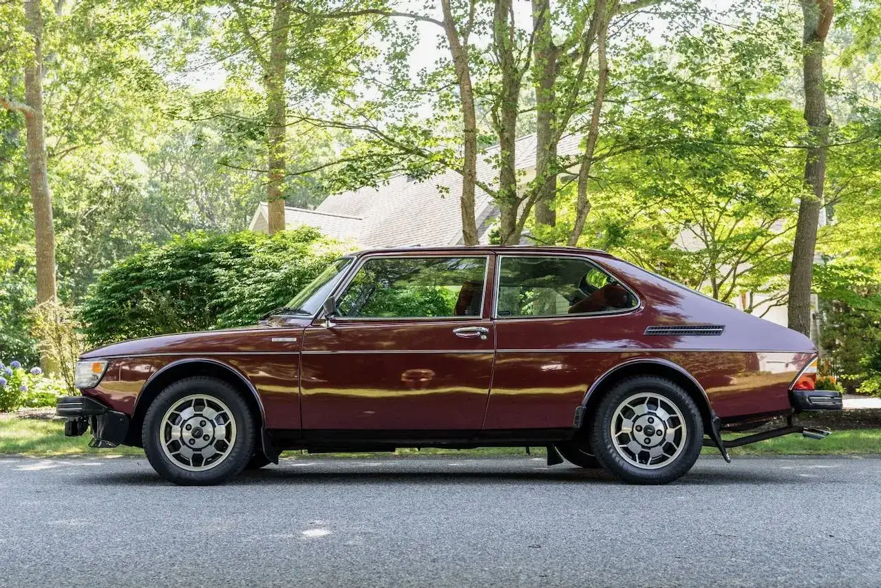 En mörkröd Saab 99 står parkerad längs en grönskande väg.