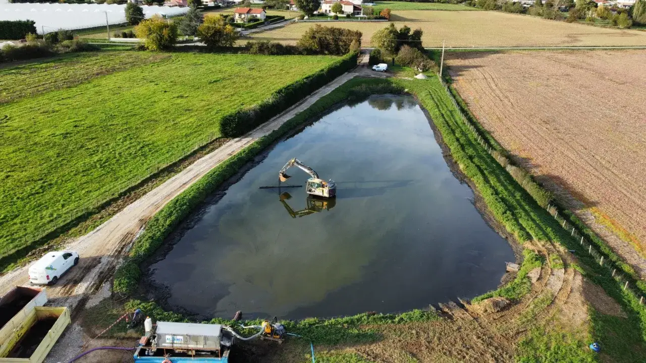 Un excavateur travaille dans un étang, entouré de champs verts et de **roseaux**. Une pompe est installée sur la rive.