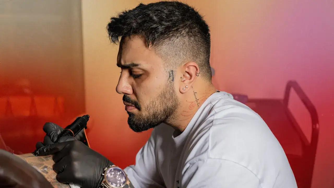 Un homme se fait tatouer discrètement le cou. Le tatoueur, ganté, utilise sa machine sur le bras du client.