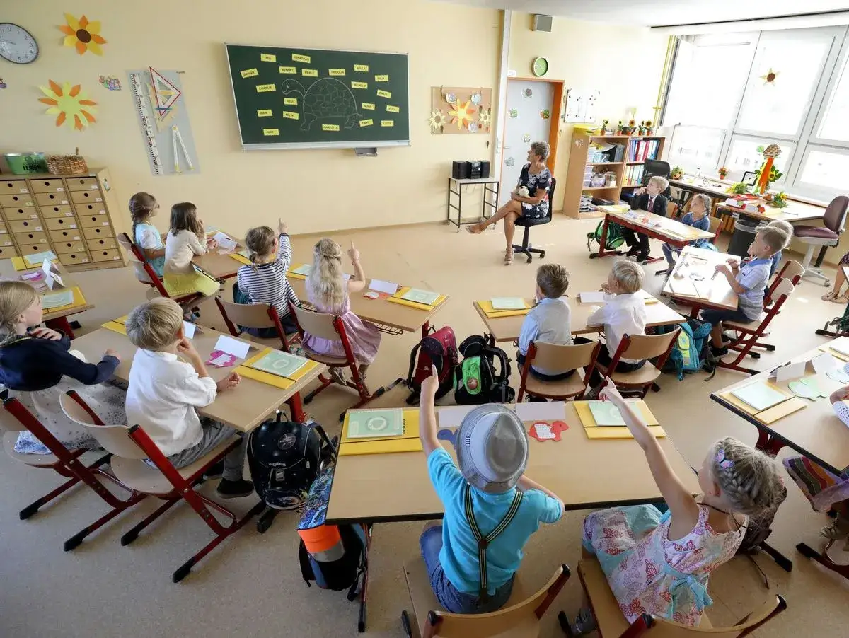 Grundschulkinder nehmen an Kennenlernspielen teil. Sie sitzen an ihren Tischen, einige heben die Hand, während die Lehrerin vorne sitzt.
