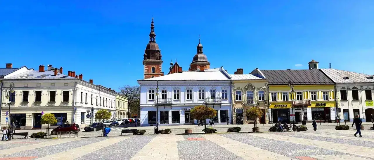 Słoneczny rynek w Nowym Sączu z zabytkowymi kamienicami i wieżami kościelnymi w tle.