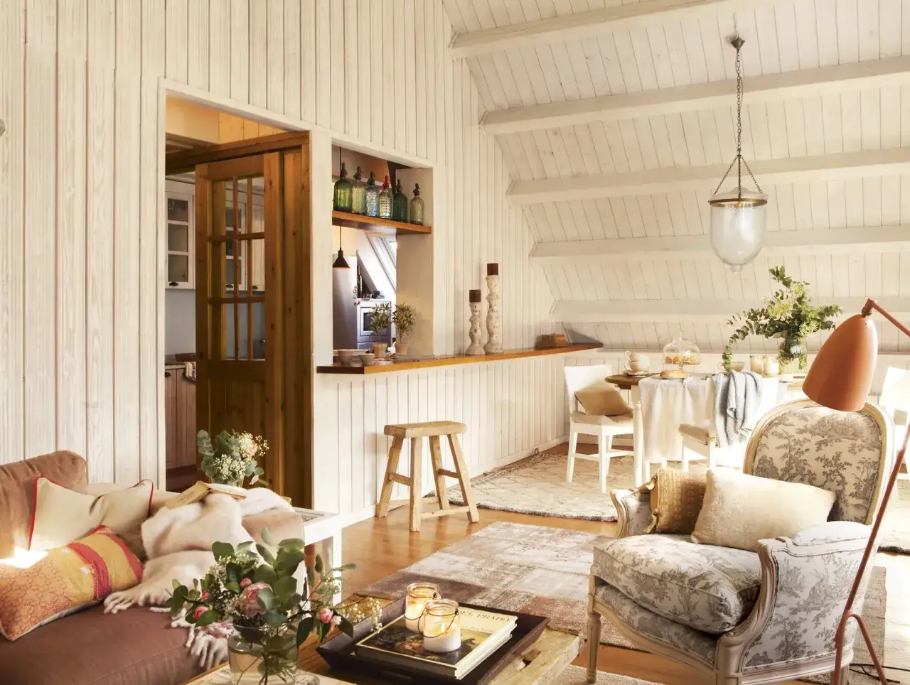 Acogedor sal&oacute;n con sof&aacute;, sill&oacute;n y mesa de comedor bajo un **techo inclinado interior** de madera.