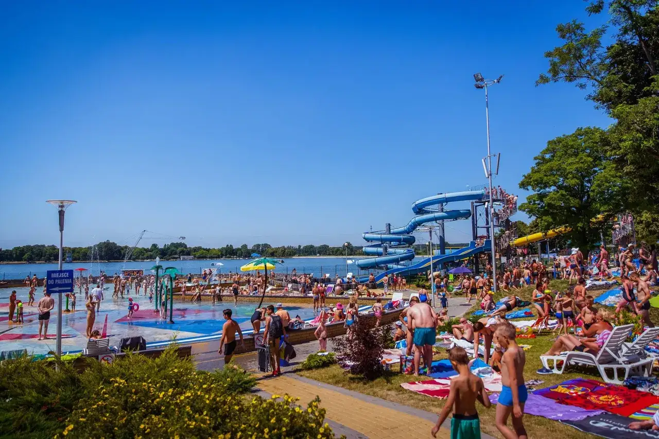 Tłumy ludzi korzystają z atrakcji wodnych na **zalewie zemborzyckim**. Widać zjeżdżalnie, brodziki i plażowiczów odpoczywających na leżakach.