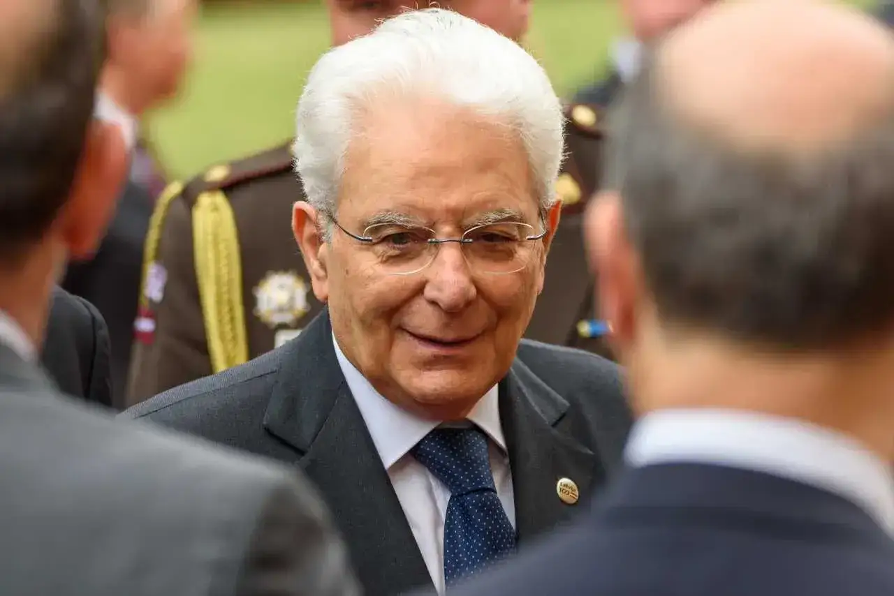 Uśmiechnięty prezydent Włoch, Sergio Mattarella, w okularach i granatowym krawacie, rozmawia z innymi mężczyznami.