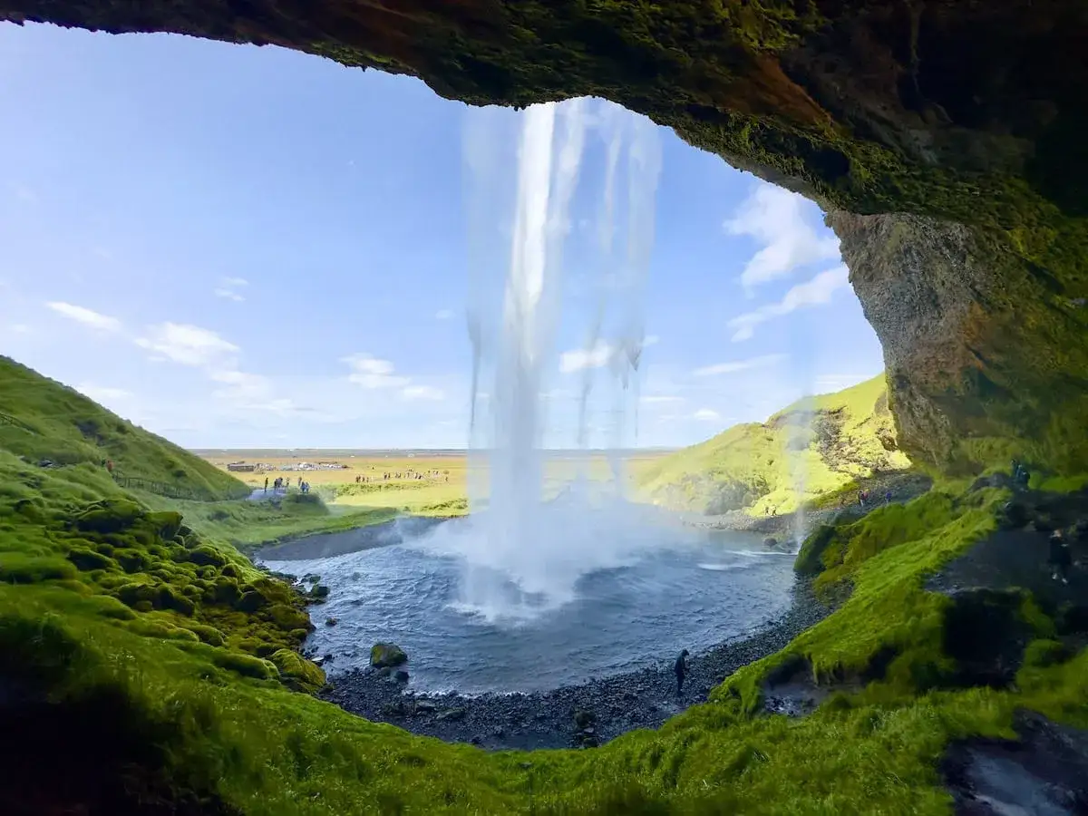 View from behind a waterfall on an Iceland Ring Road itinerary. Lush green moss covers the rocks, with a powerful cascade plunging into a pool below.