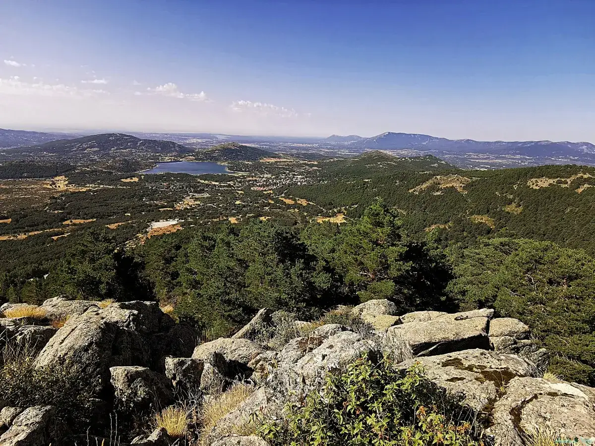 Vista panorámica del **valle de la barranca** con un lago azul y montañas verdes bajo un cielo despejado. Rocas en primer plano.