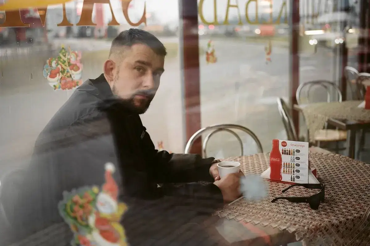 Mężczyzna z brodą siedzi przy stoliku, trzymając kubek kawy. Na stole leży menu z butelką Coca-Coli. W tle widać świąteczne ozdoby.