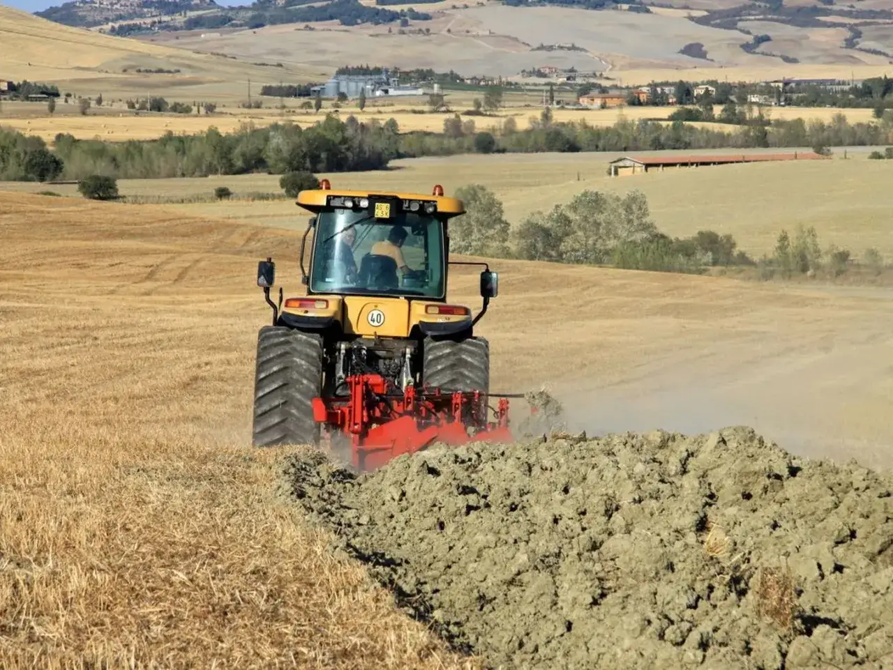 Trattore giallo in primo piano esegue l'aratura del terreno, sollevando terra e polvere in un paesaggio rurale collinare.
