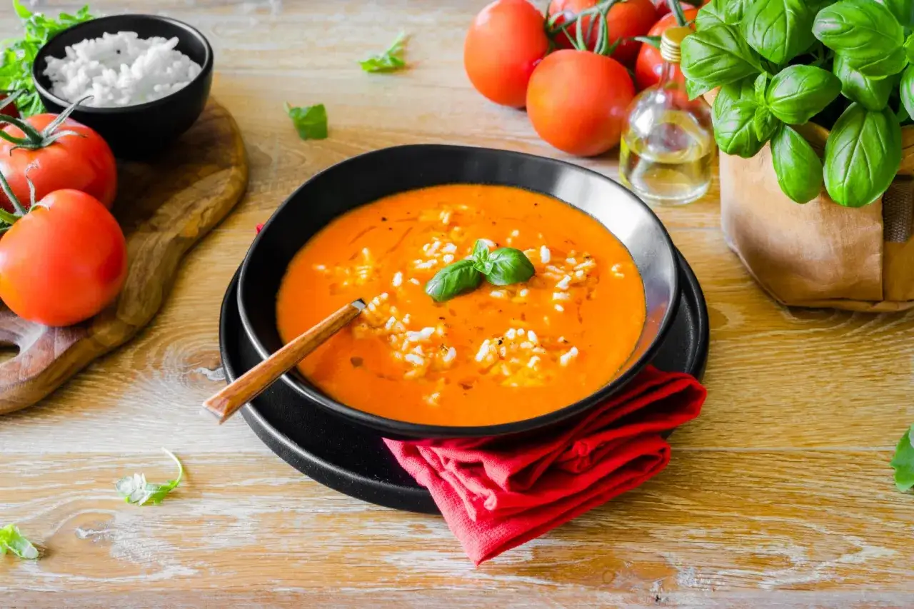Eine Sch&uuml;ssel mit leuchtend orangefarbener Tomatensuppe, verfeinert mit Reis und Basilikum. Frische Tomaten und ein Kr&auml;utertopf vervollst&auml;ndigen das Bild des k&ouml;stlichen sugo di pomodoro.