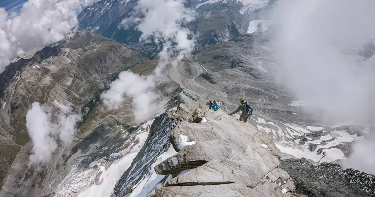 Wspinacze na skalistej grani, w tle majestatyczne góry i lodowce. W oddali widać miasteczko. To trudne wejście na Matterhorn.