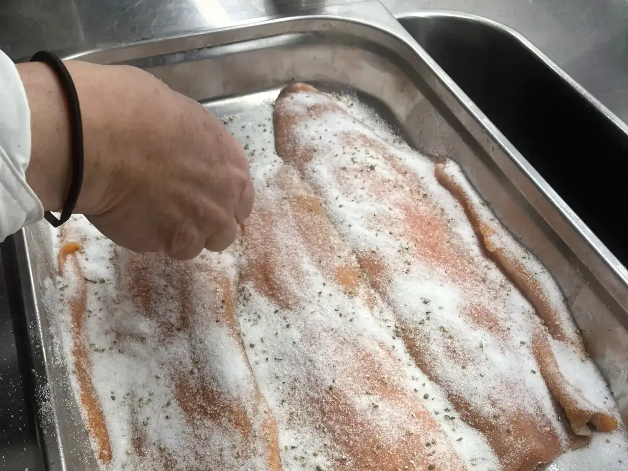 En hand strör salt och peppar över filéer av gravad regnbåge i en metallform.
