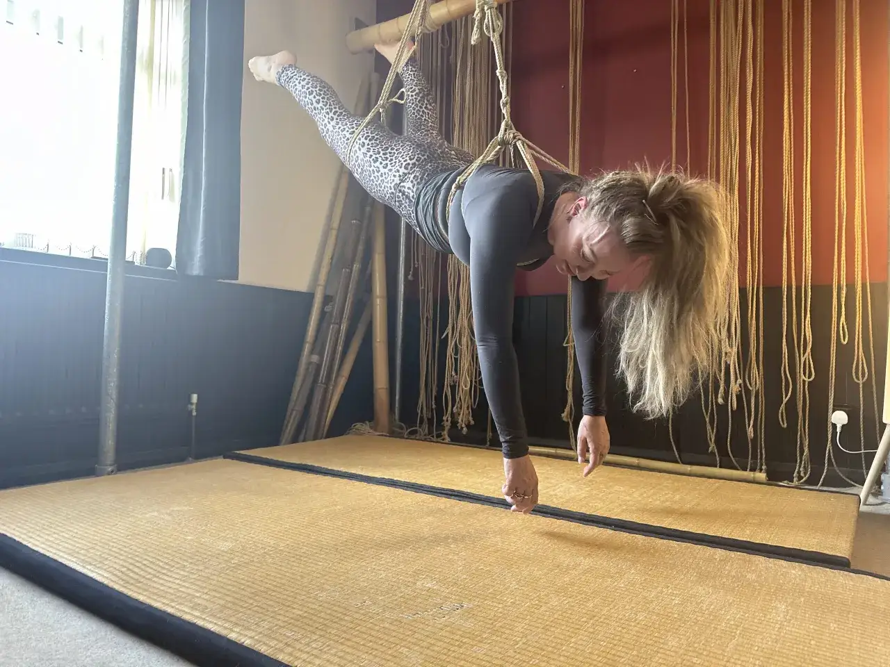 Kobieta w panterkowych legginsach wisi na linach, demonstrując podstawy shibari. Jej ciało jest napięte, a dłonie wyciągnięte w dół.
