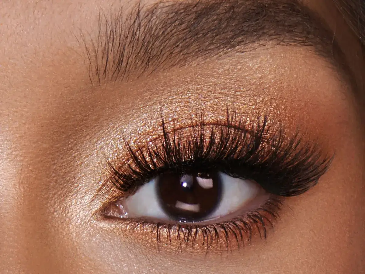 Un œil marron sublimé par un maquillage doré scintillant et des cils longs et volumineux.
