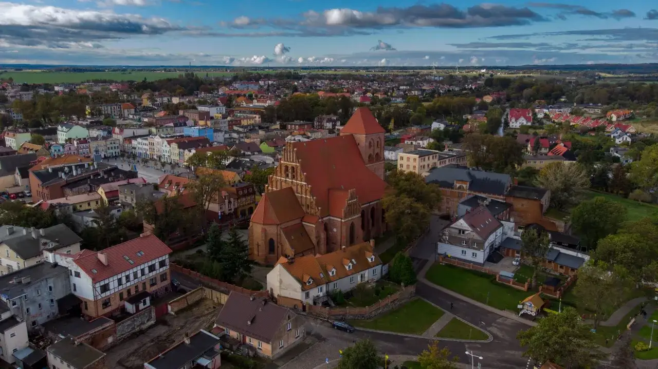 Widok z lotu ptaka na miasto z dominującym ceglanym kościołem w Pucku. Dachy domów i zieleń tworzą malowniczy krajobraz.