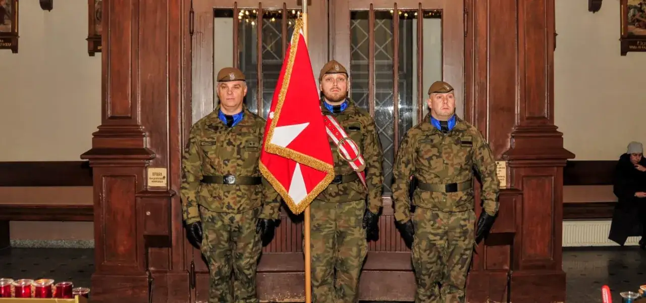 Trzech żołnierzy w mundurach stoi przed drzwiami kościoła, jeden z nich trzyma flagę. Nie widać, czy żołnierz klęka w kościele.