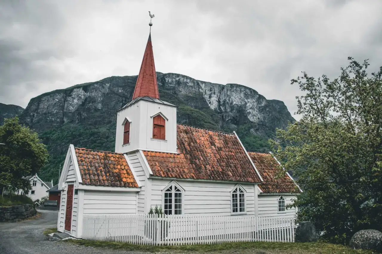 Biały kości&oacute;łek z czerwoną wieżą i dachem, otoczony białym płotkiem, na tle surowych g&oacute;r. Symbol norweskiej religii i spokoju.