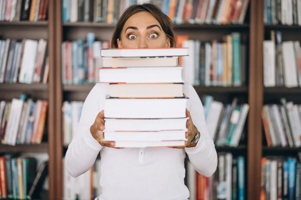 Zaskoczony bibliotekarz z wielką stertą książek w rękach, otoczony regałami pełnymi literatury.
