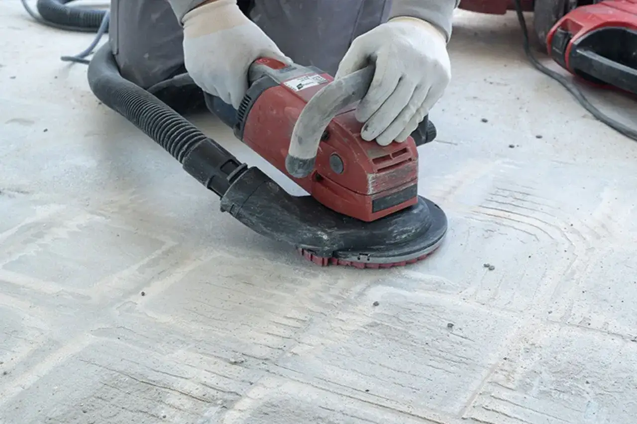 Gants blancs et ponceuse rouge en action pour poncer du béton. Le tuyau d'aspiration est connecté à la machine.