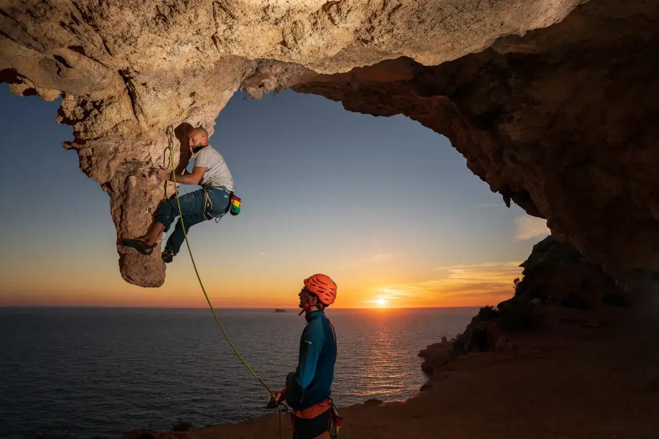 Ein Kletterer genie&szlig;t das Malta Klettern in einer H&ouml;hle bei Sonnenuntergang. Ein zweiter Kletterer sichert ihn.
