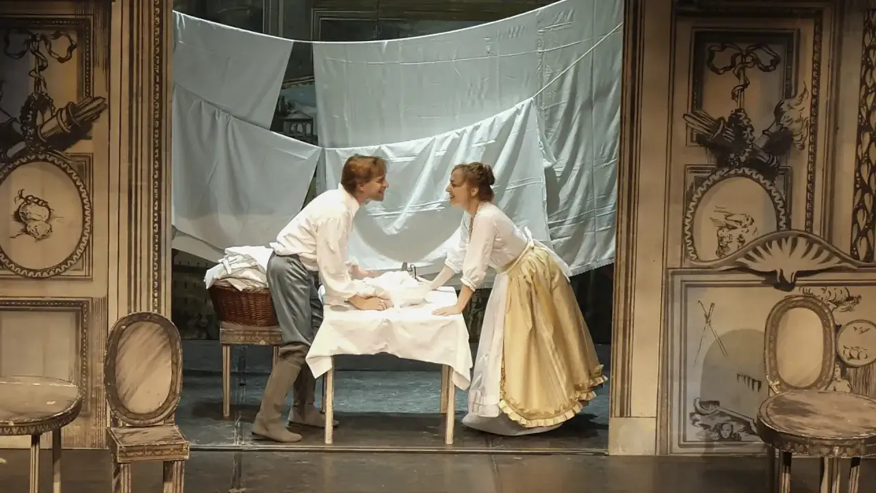Scena teatrale con due attori che interagiscono attorno a un tavolo coperto da un lenzuolo bianco. Le recensioni per