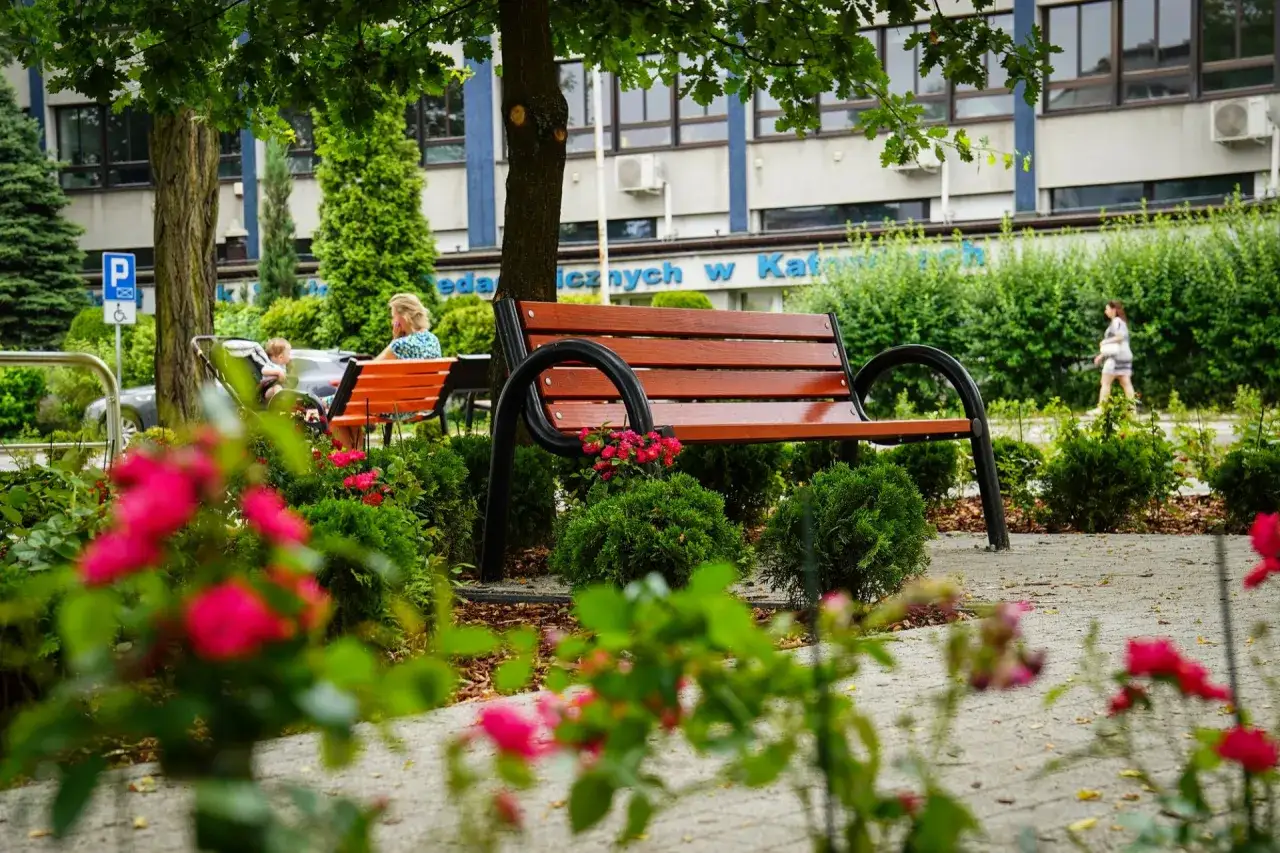Piękny teren zieleni urządzonej z ławkami, krzewami i kwitnącymi r&oacute;żami. Kobieta z dzieckiem siedzi na ławce, a w tle przechodzi inna osoba.