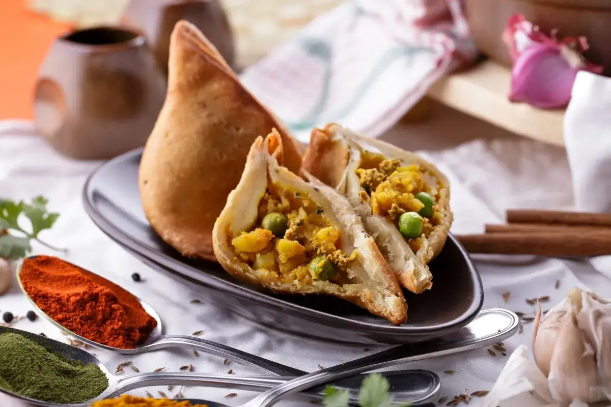 Chrupiący indyjski pierożek samosa, przekrojony na pół, odsłaniający aromatyczne nadzienie z ziemniaków i groszku. Obok przyprawy: papryka, curry, czosnek.