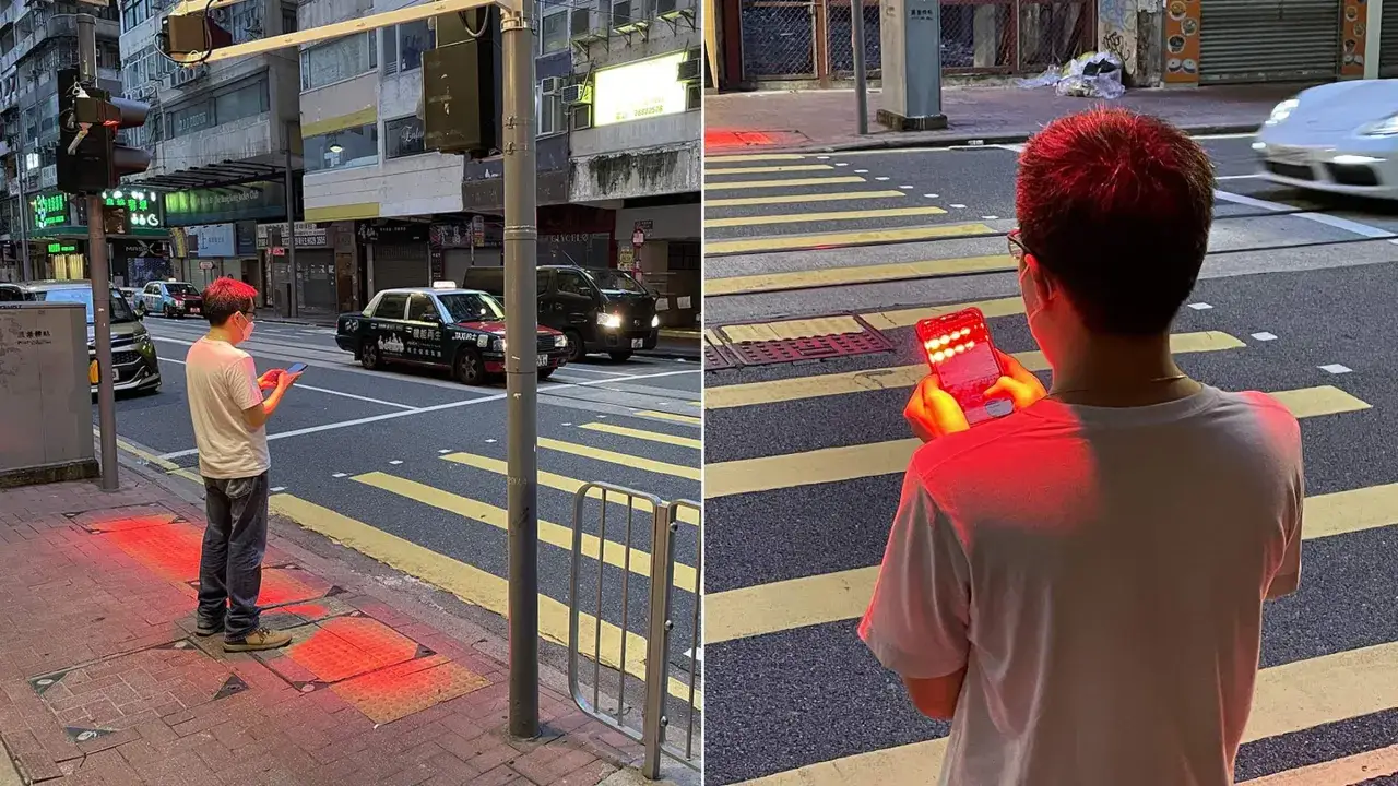 Un homme attend au feu rouge, son t&eacute;l&eacute;phone &eacute;clairant son visage d'une lueur rouge. Des lumi&egrave;res rouges se refl&egrave;tent sur le trottoir.